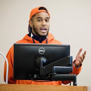 Male in orange hat and sweatshirt speaking from behind a computer screen